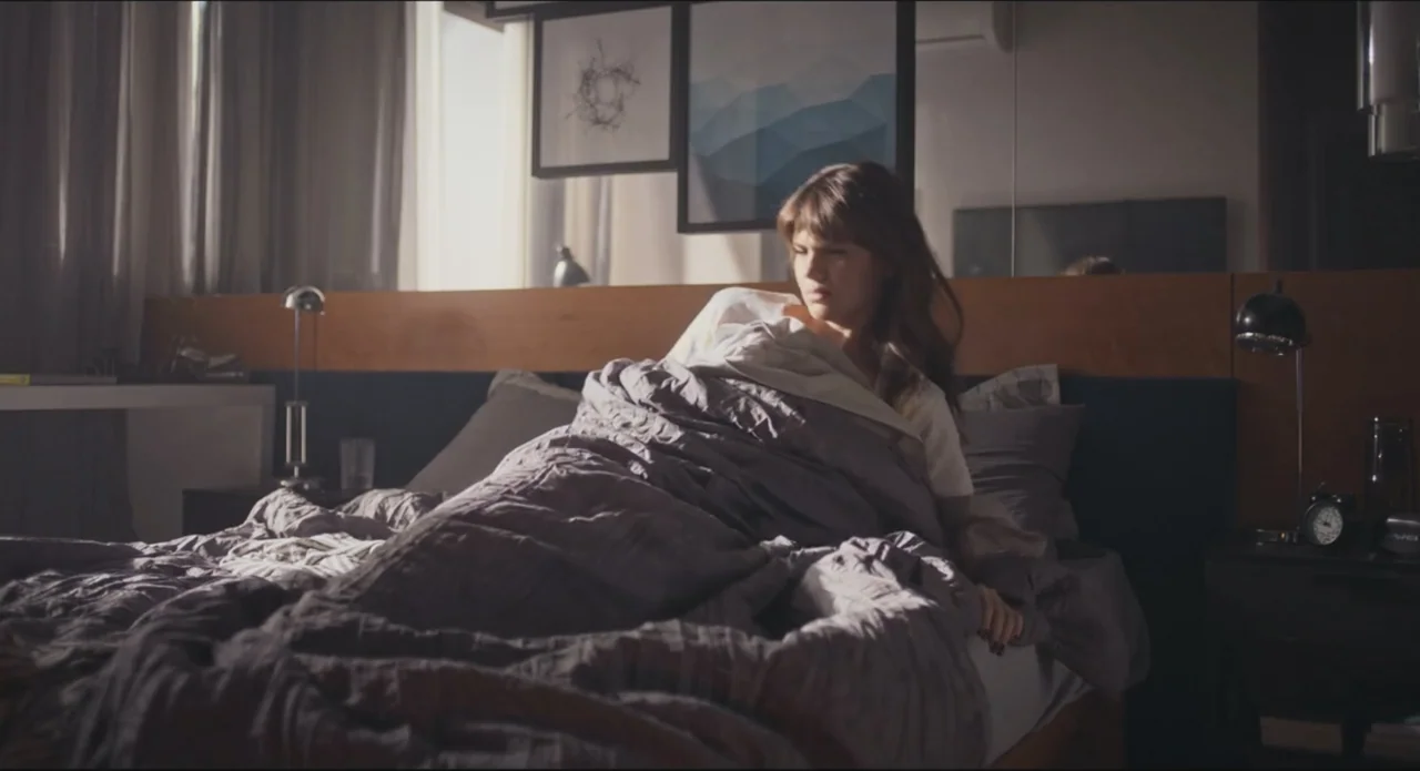 Proof of Love (2022) movie still: medium — A modern room features a bed with gray linens and abstract art on walls. A young woman in a white t-shirt…
