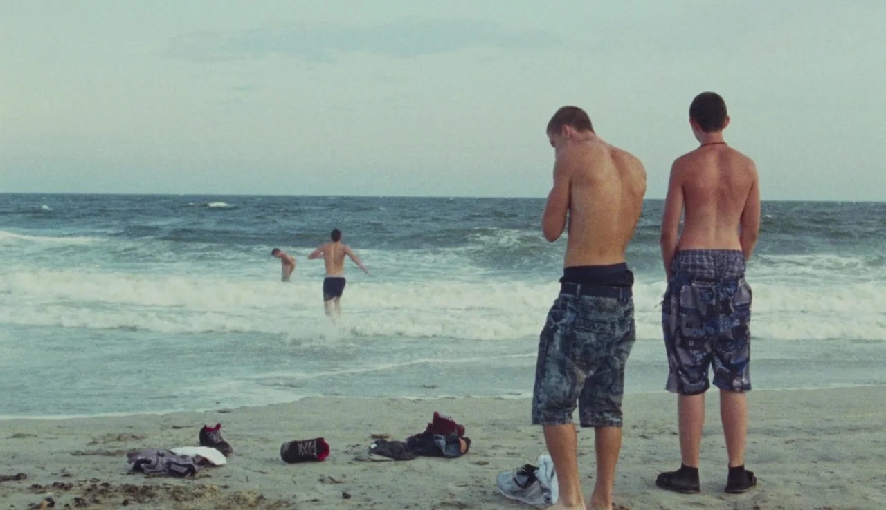 Beach Rats (2017) movie still: fullbody — A sandy shore overlooks a gray, overcast ocean with light surf conditions. Two young men stand in the…