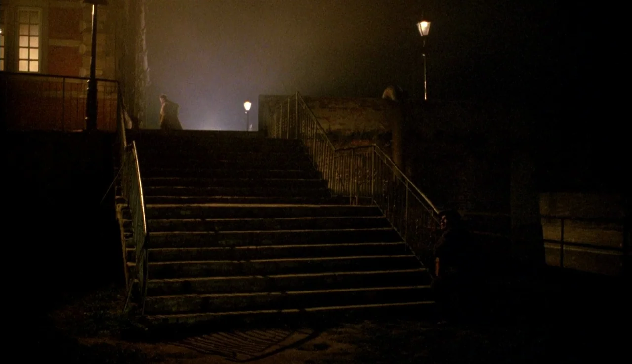 The Rainbow Thief (1990) movie still: wide — A set of dark stone stairs connects to a building illuminated by warm lamp light. Two men are present,…