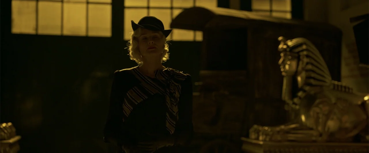Marlowe (2023) movie still: medium — A dark room contains an ancient golden statue and a large wooden crate. A woman wearing a 1930s style hat and…