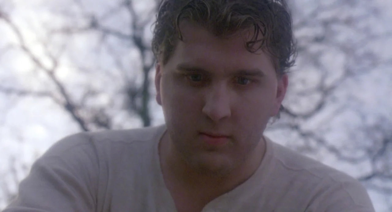 River’s Edge (1986) movie still: closeup — An outdoor area with bare, leafless trees against an overcast white sky. A young man stares downward at…