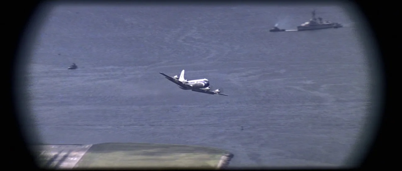 Moonraker (1979) movie still: wide — A vintage propeller airplane flies over a vast expanse of open dark blue water. A large naval vessel sits in…