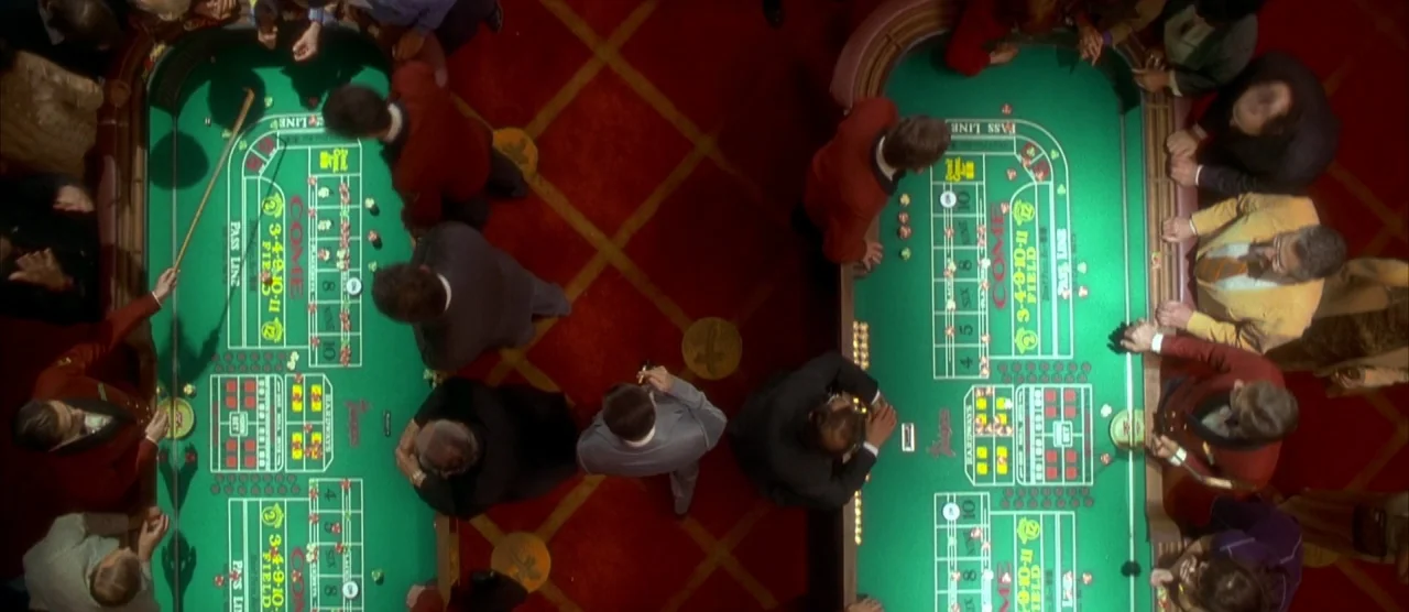 Casino (1995) movie still: wide — An aerial view of a crowded casino floor features two long green craps tables. People gather around the tables to…