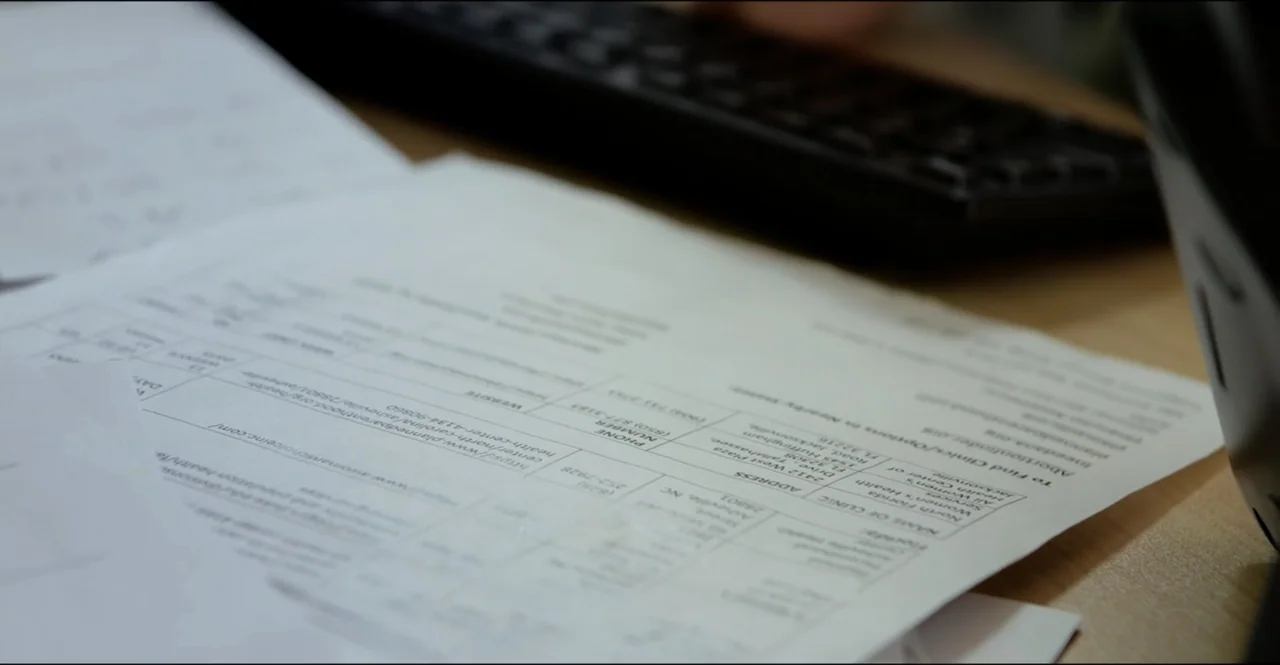 The Devil Is Busy (2024) movie still: closeup — A close-up view of a printed informational document resting on a wooden desk. A blurred computer…