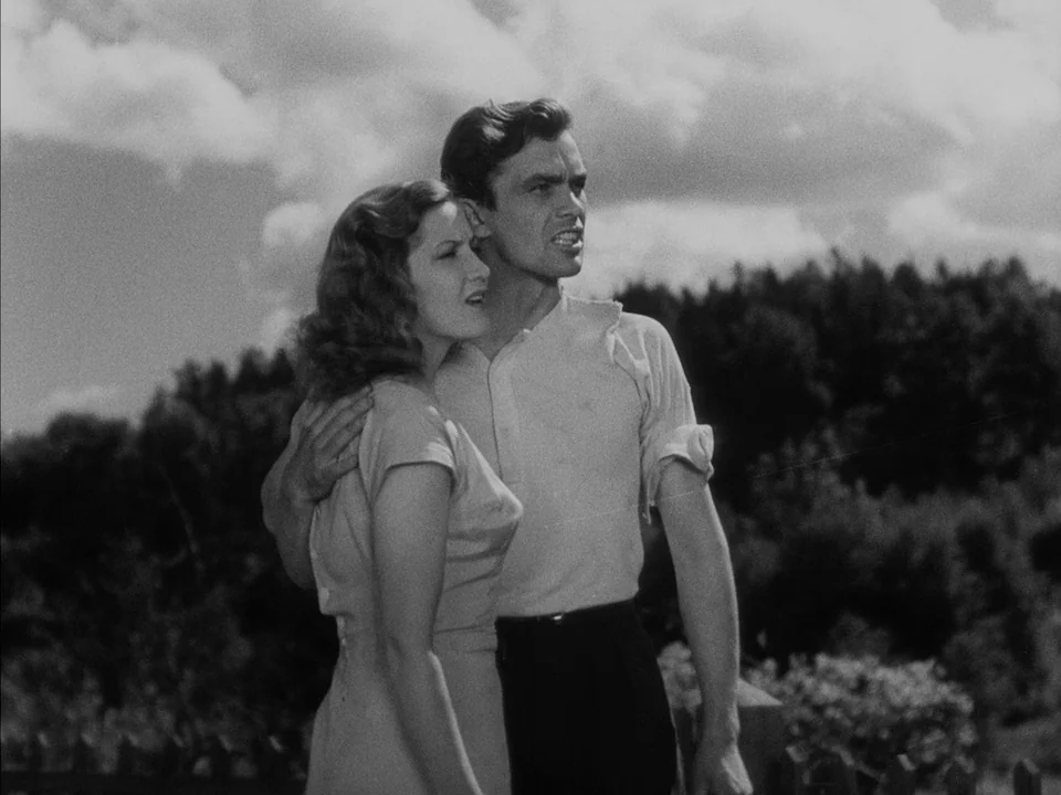 It Rains On Our Love (1946) movie still: medium — A man and woman stand together in a grassy field with trees visible behind. The man holds the…