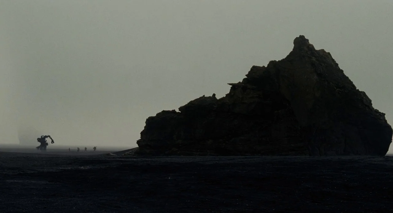 Noah (2014) movie still: wide — A vast black sand beach stretches beneath a thick, overcast, monochromatic gray sky. A large, jagged volcanic rock…