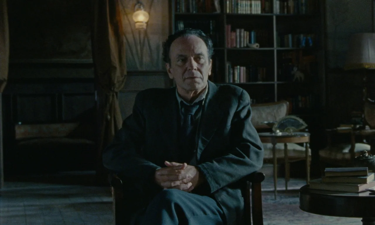 Close Your Eyes (2023) movie still: medium — A dark study features a tall bookcase and a small table with a lamp. A man sits in a chair with his…