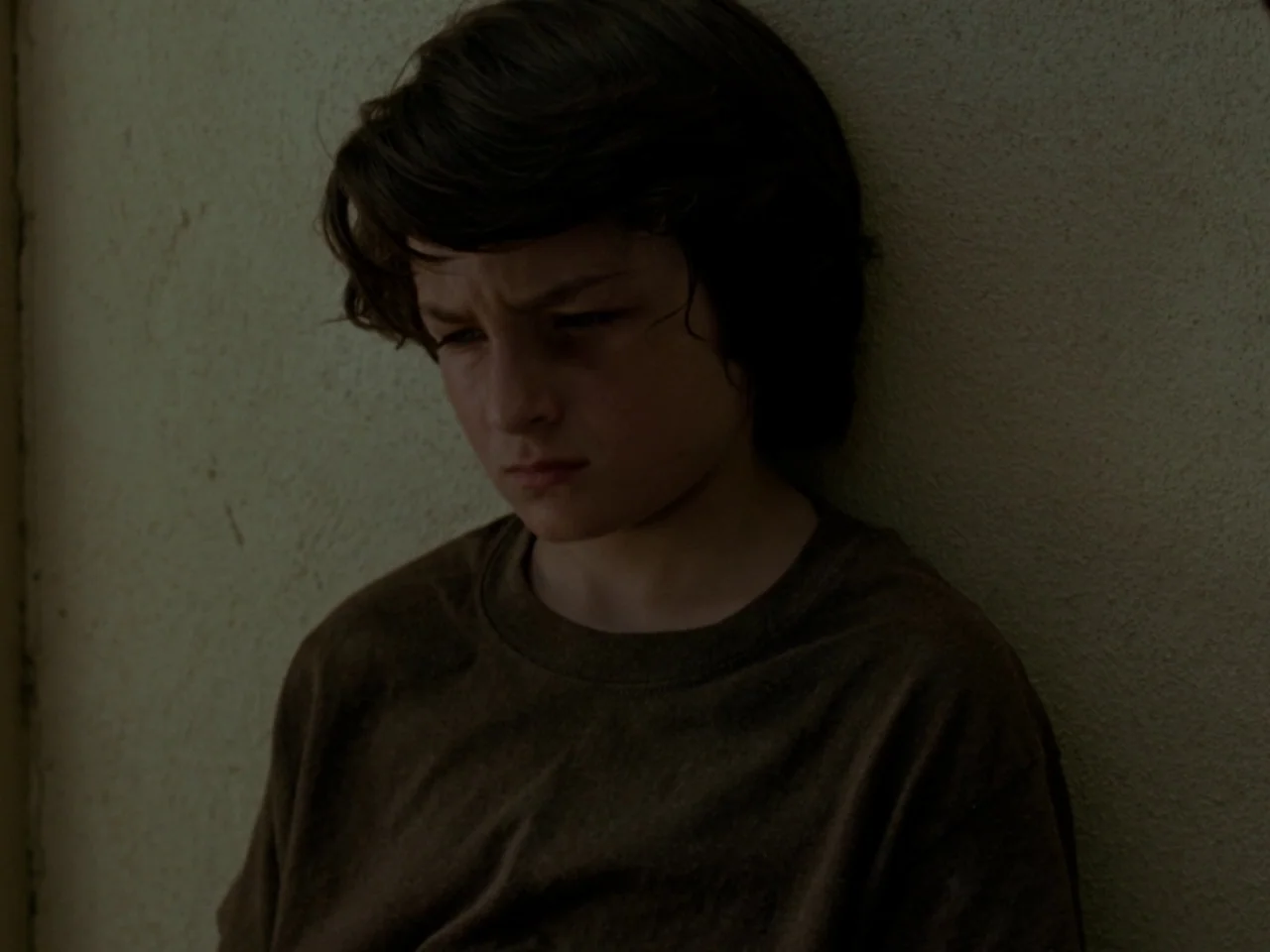 Mid90s (2018) movie still: closeup — A young boy stands in front of a plain, textured beige wall. He leans his body against the surface while his…