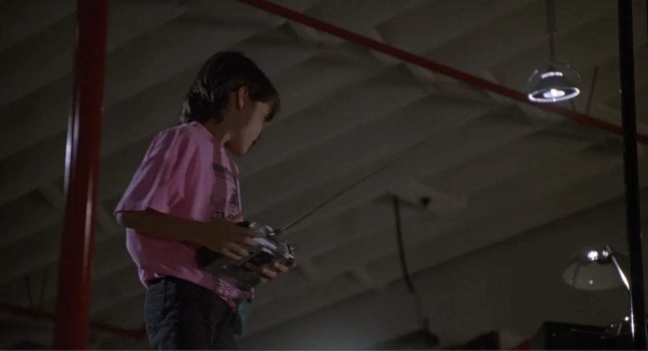 F/X2 (1991) movie still: medium — An interior garage space with exposed ceiling beams and hanging pendant lights. A young boy holding a radio…