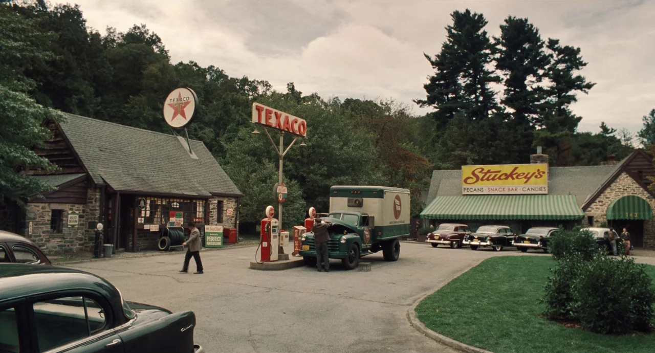 The Irishman (2019) movie still: wide — A 1950s roadside stop features a stone gas station and a nearby snack bar. Two men attend to vehicles on the…