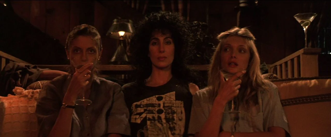 The Witches of Eastwick (1987) movie still: medium — Three women sit together on a plush sofa in a dimly lit room at night. They each hold a martini…