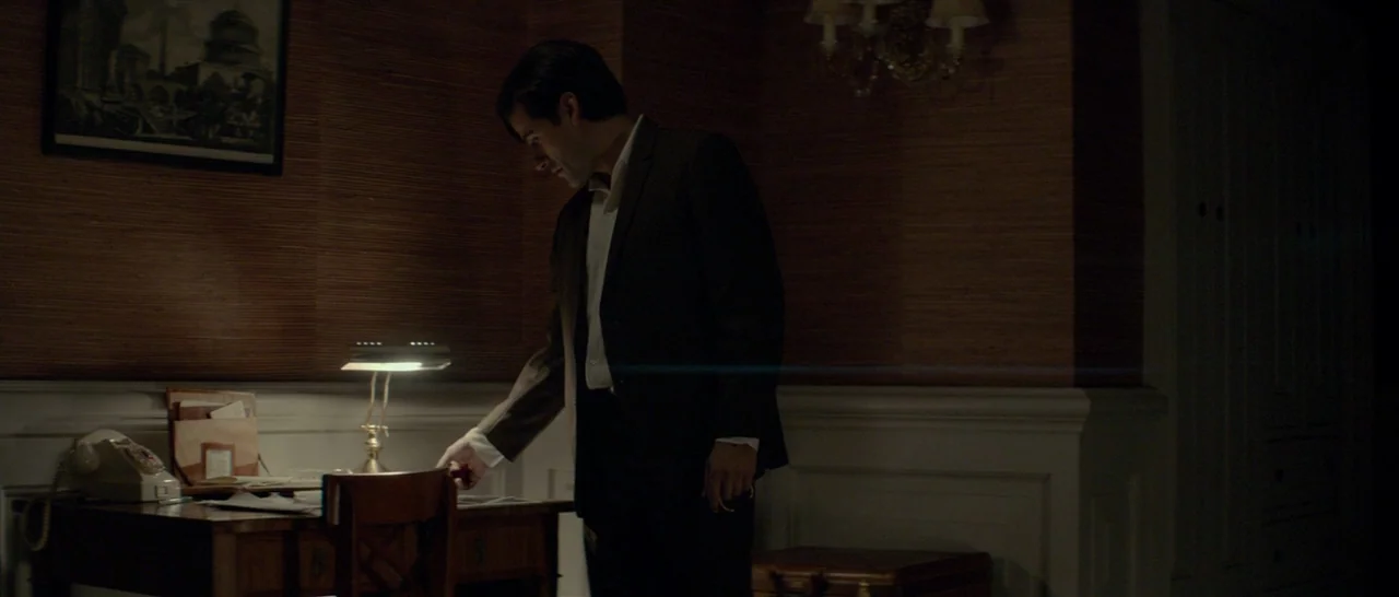 The Two Faces of January (2014) movie still: medium — A dimly lit room features wood-paneled walls and a classic desk with a lamp. The man stands by…