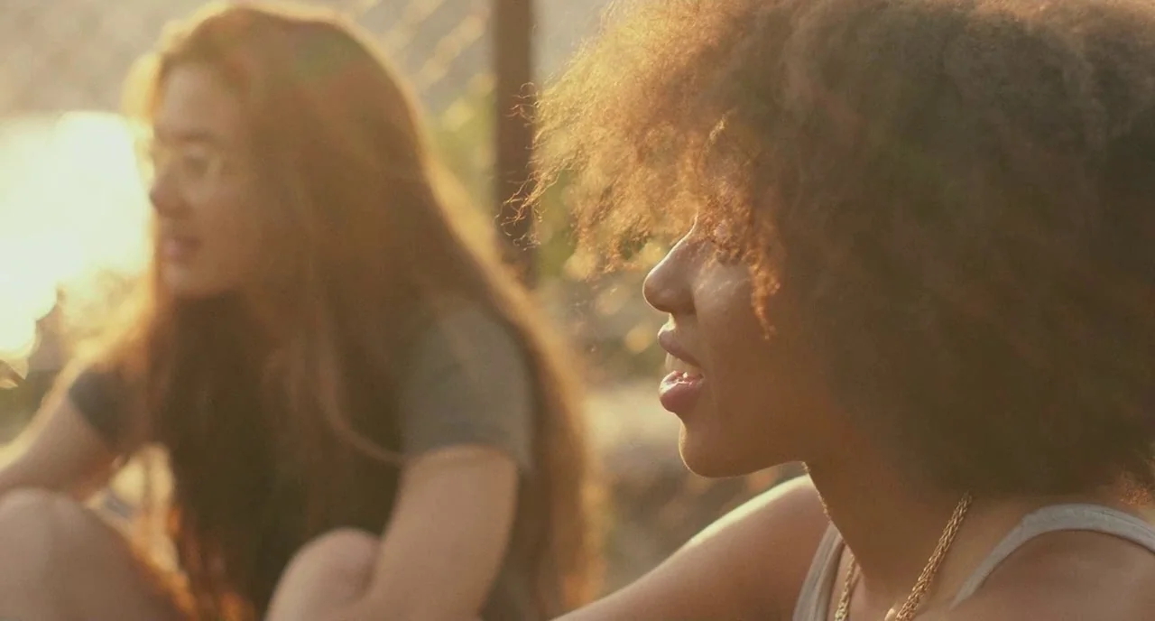 Skate Kitchen (2018) movie still: medium — Two young women sit outdoors near a chain link fence during late afternoon. They are positioned…