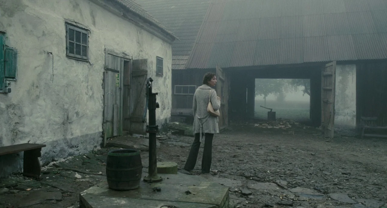 Revanche (2008) movie still: fullbody — A rural courtyard features a whitewashed building and an open-fronted wooden barn. A woman stands on the…