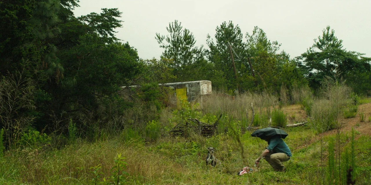 The Death of Dick Long (2019) movie still: wide — An overgrown, grassy field contains a dilapidated structure with a prominent yellow door. A man…