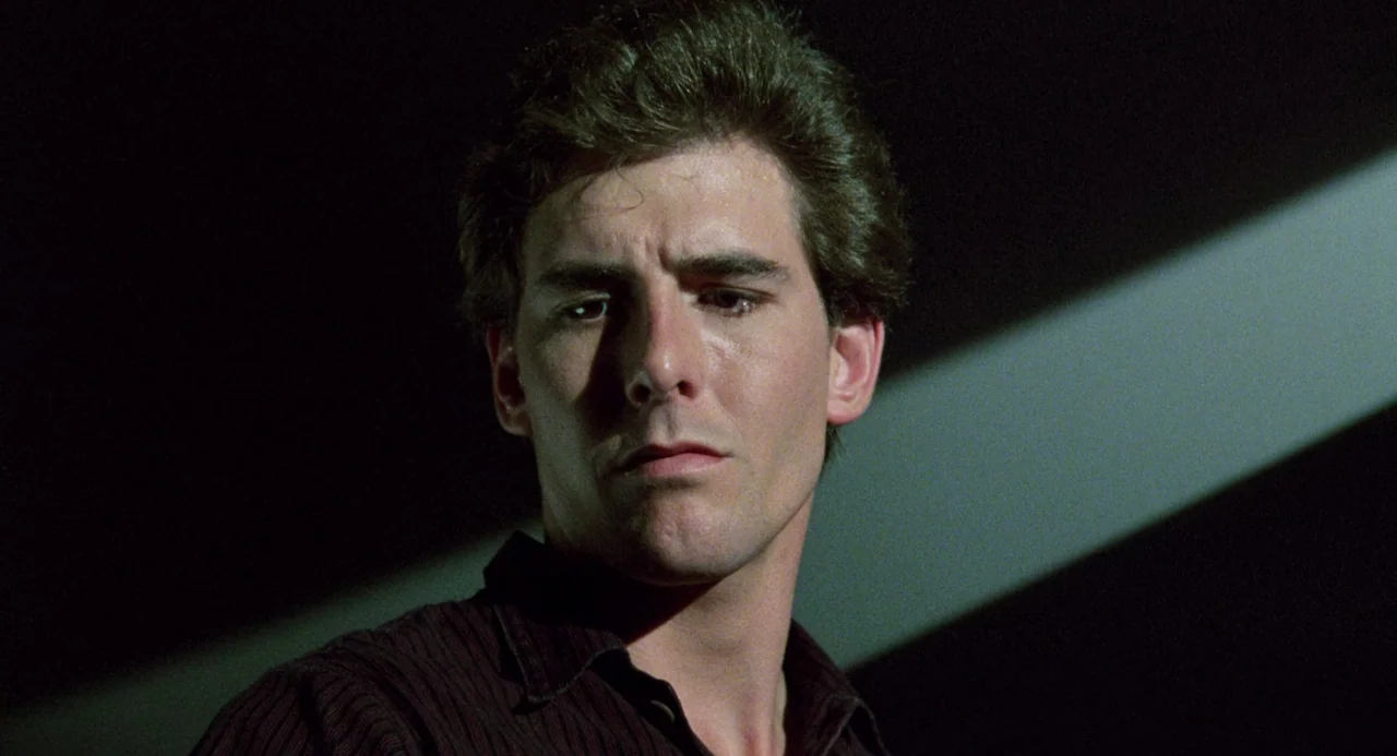 Crimes of Passion (1984) movie still: closeup — A man stands in a dimly lit environment with a diagonal beam of light. He faces to the side while…