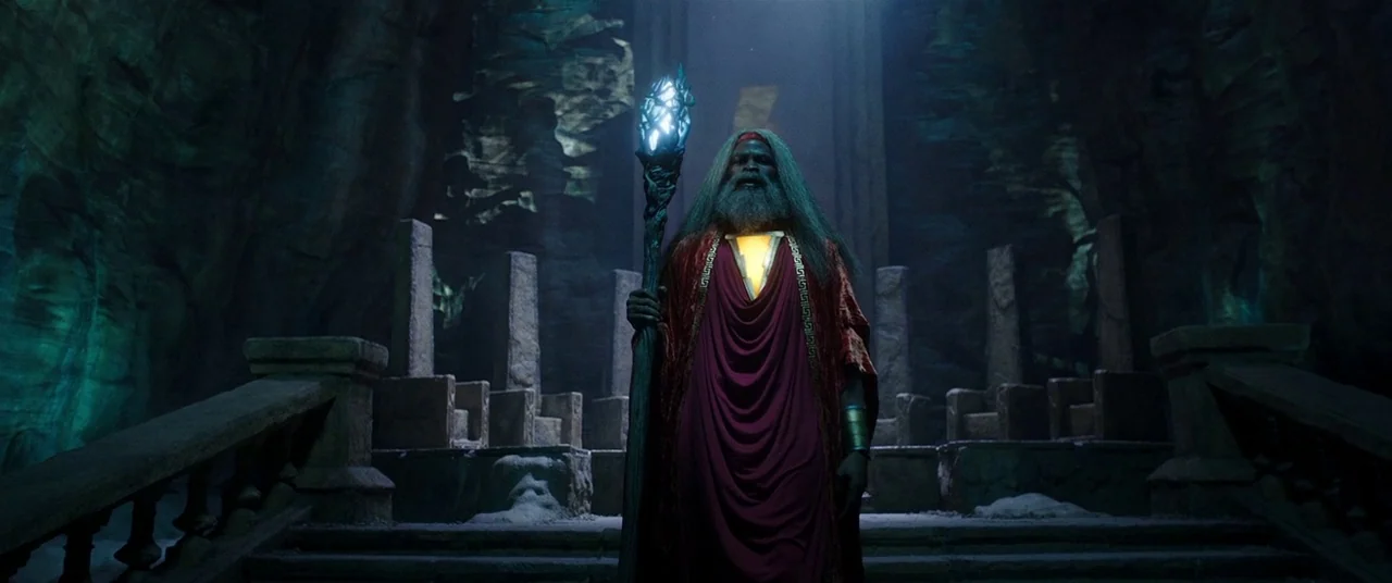 Shazam! (2019) movie still: fullbody — An elderly man in robes stands within a dark, ancient-looking cavernous hall made of stone. The man holds a…