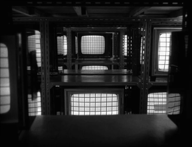 Alphaville (1965) movie still: establishing — A dimly lit storage room features multiple shelves filled with vintage television sets. Each screen…