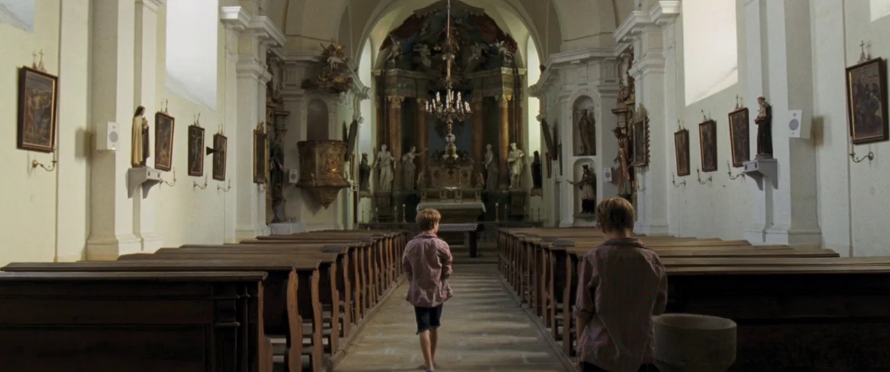 Goodnight Mommy (2014) movie still: wide — A historic stone church interior features rows of wooden pews and religious statues in niches. Two boys…