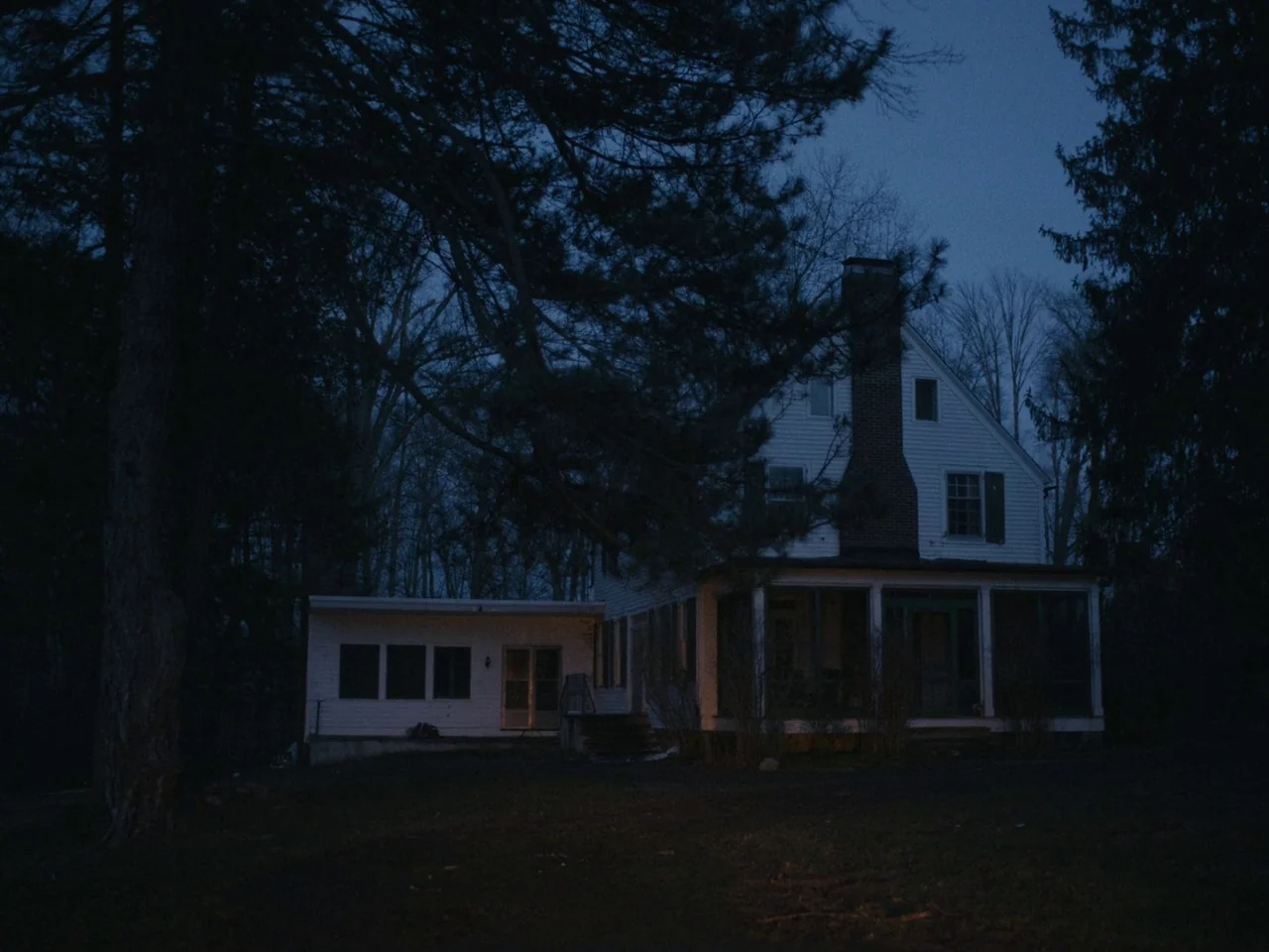 I’m Thinking of Ending Things (2020) movie still: establishing — A two-story white clapboard house with a brick chimney stands among tall pine…