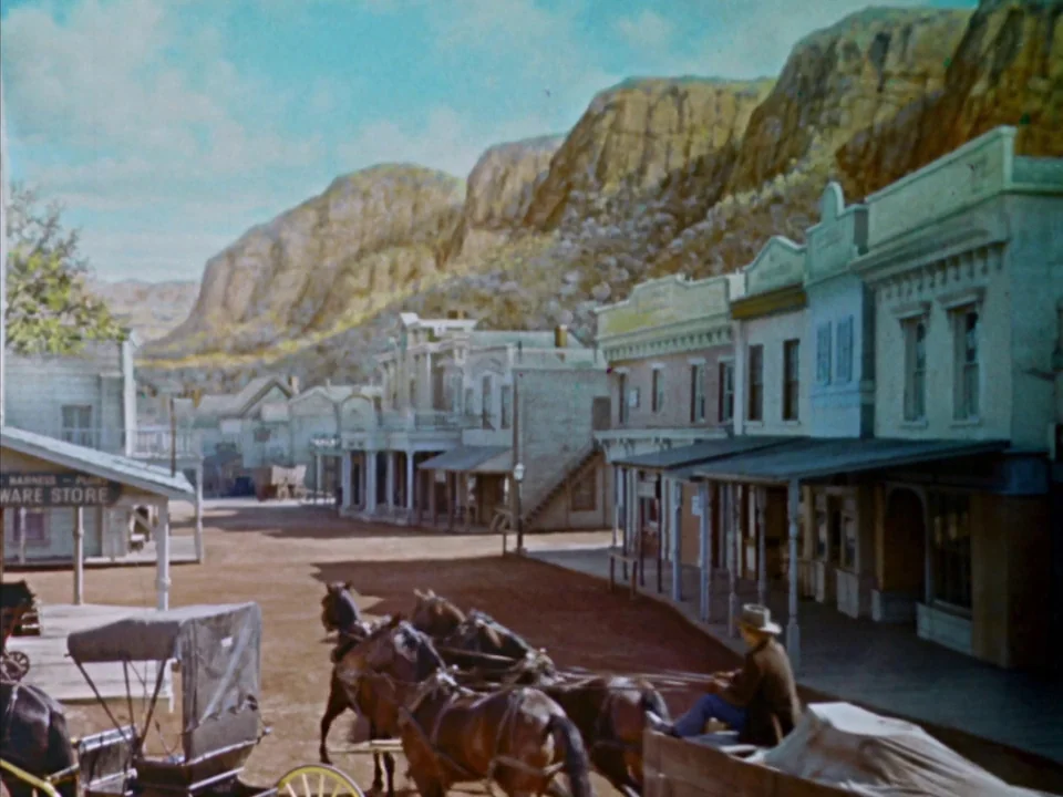 Johnny Guitar (1954) movie still: wide — A dusty main street in a 19th-century frontier town lined with two-story wooden buildings. A man sits on a…