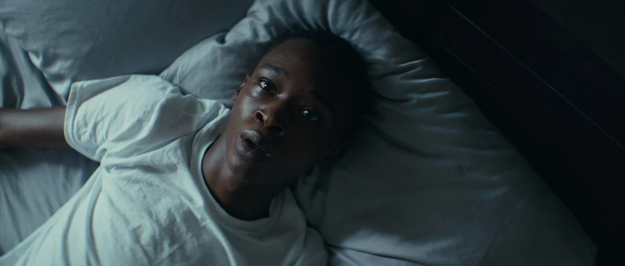 Moonlight (2016) movie still: medium — A person lies on a bed with light-colored sheets in a dark room. The subject looks directly toward the…