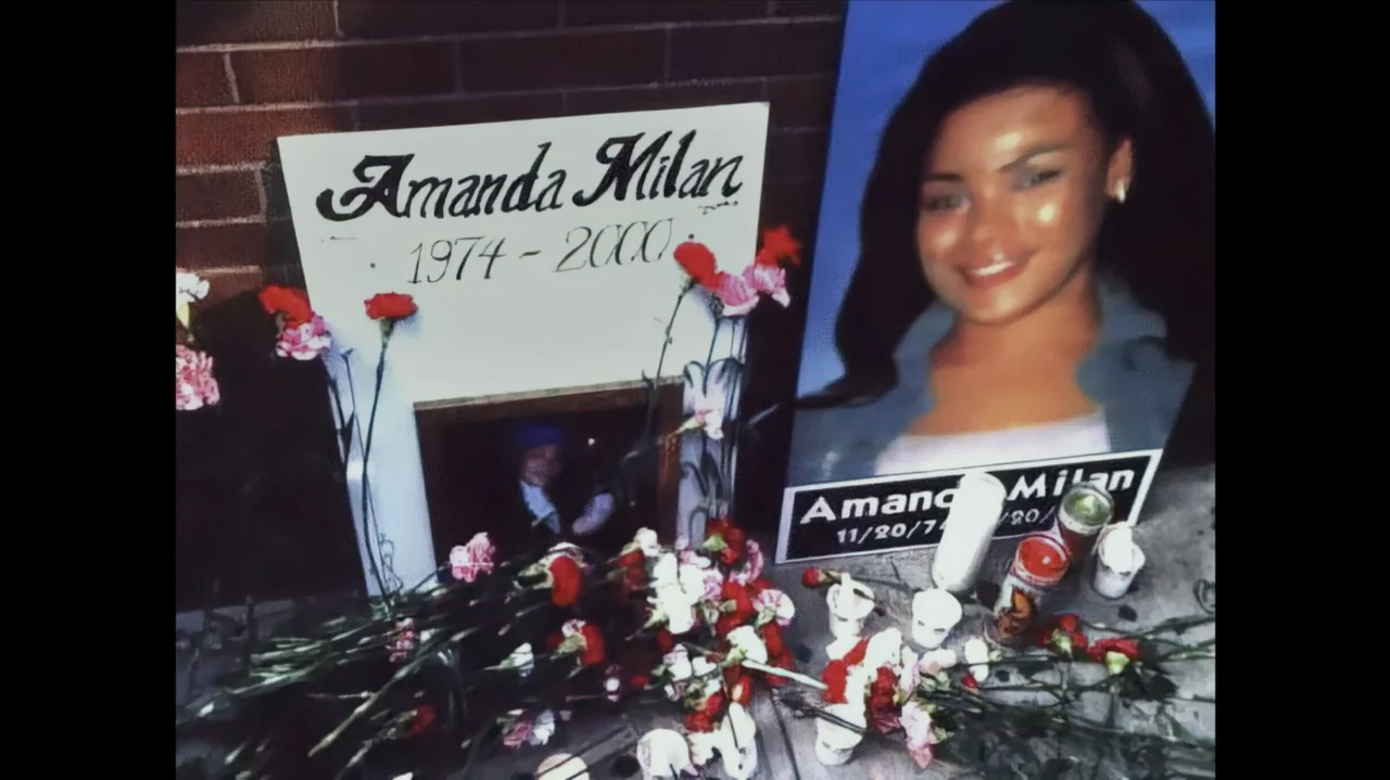 The Stroll (2023) movie still: medium — A memorial display sits against a red brick wall featuring photographs and signs. Red carnations and white…