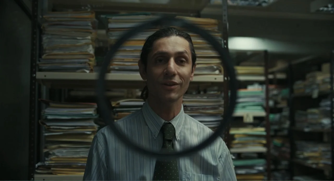 Belén (2025) movie still: medium — An archive room is filled with metal shelving units stacked high with files and papers. A man in a dress shirt…