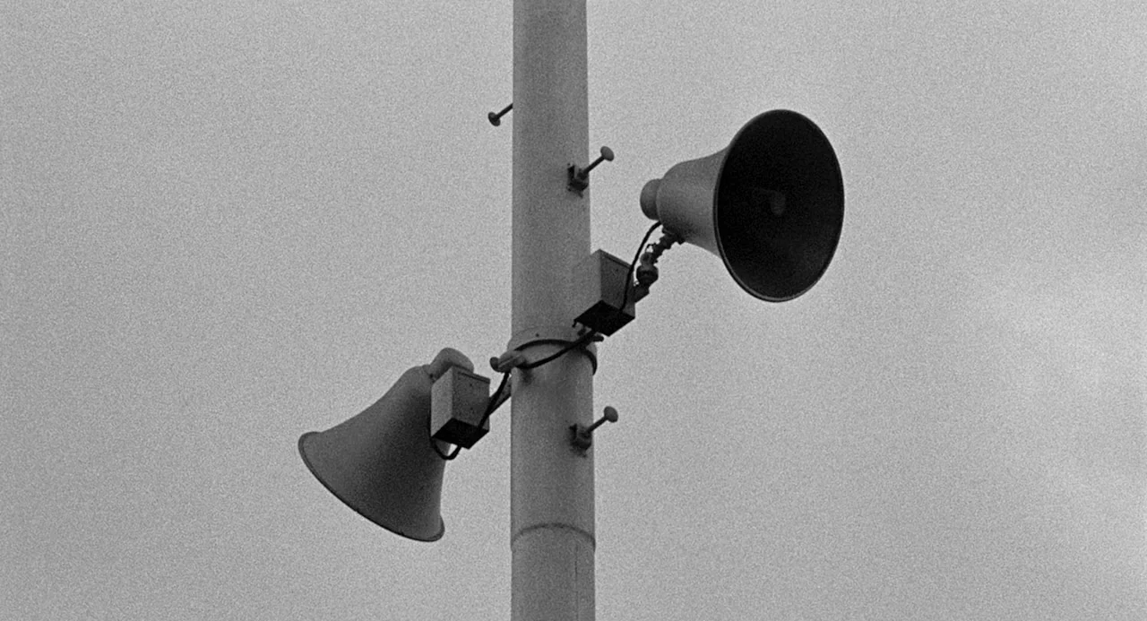 Murder By Contract (1958) movie still: closeup — A vertical utility pole stands against a featureless overcast sky. Two industrial horn speakers are…
