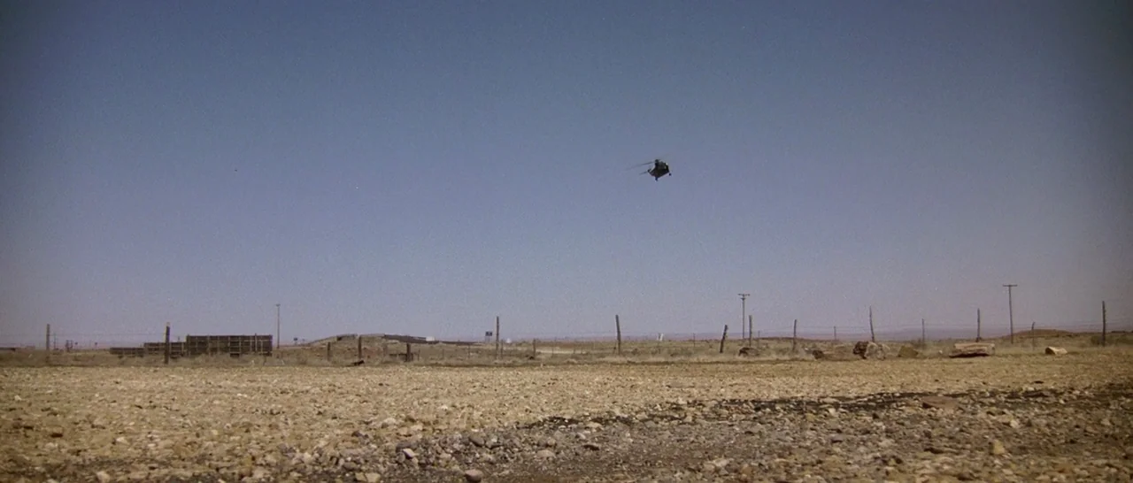 Starman (1984) movie still: wide — An arid, rocky desert plain stretches under a clear, bright blue sky. A distant small helicopter flies…