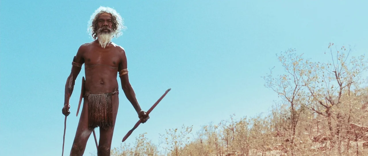 Australia (2008) movie still: fullbody — An arid, sun-drenched landscape with scrubby trees and dry earth under a clear sky. A man stands in the…
