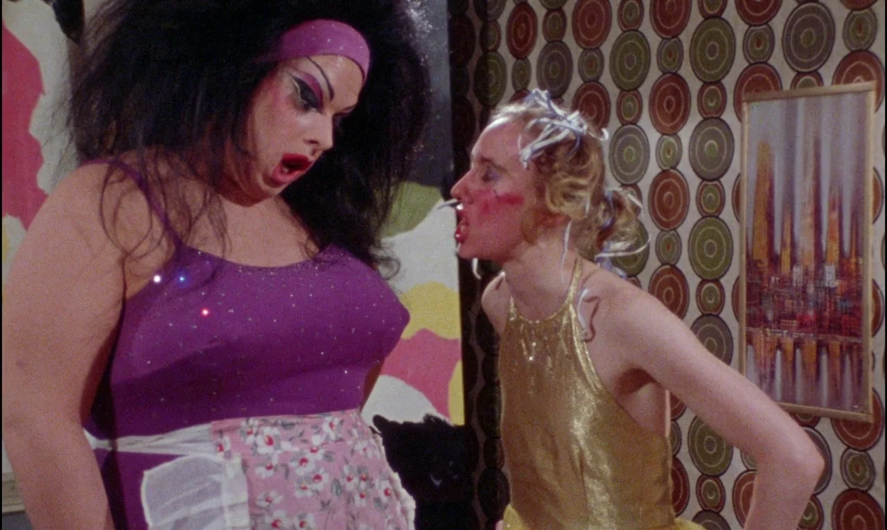 Female Trouble (1974) movie still: medium — A 1970s living room features patterned geometric wallpaper and a gold-framed print. Two women face each…