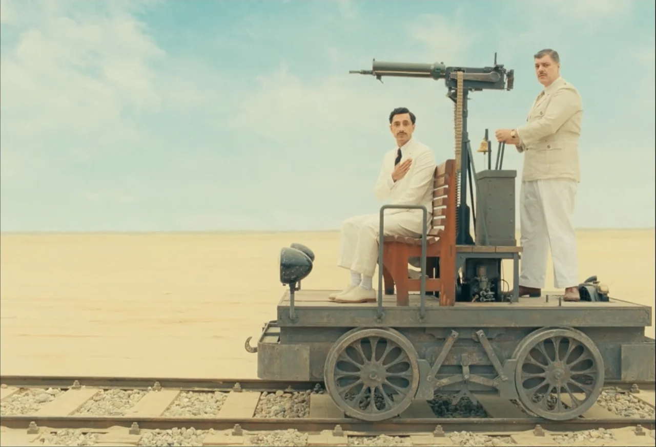 The Phoenician Scheme (2025) movie still: fullbody — A flat desert landscape stretches under a pale sky behind a metal railway cart. Two men in…
