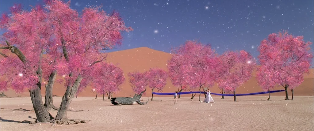 The Cell (2000) movie still: wide — A vast desert landscape features numerous trees with vibrant pink blossoms growing from sand. A woman in a white…