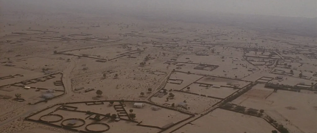 Code 46 movie still: establishing — An aerial view overlooks a vast arid settlement featuring scattered walled compounds in sand. Sparse vegetation…