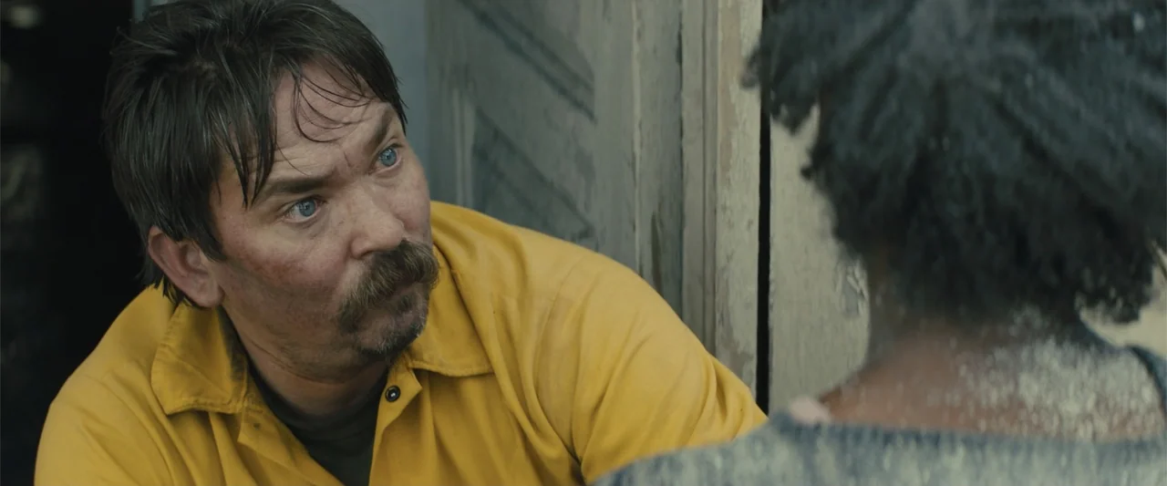 Those Who Wish Me Dead (2021) movie still: medium — A man in a yellow shirt stands near a corner in an indoor room. The man looks toward an…