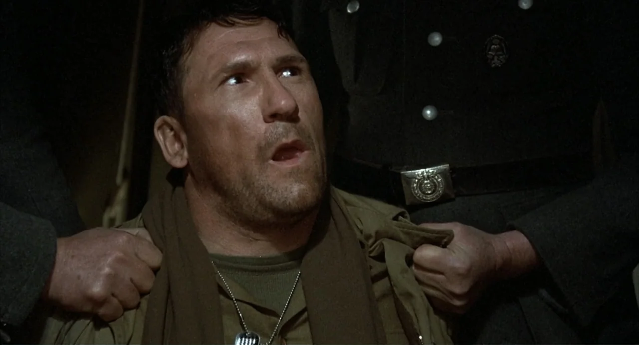 Zone Troopers (1985) movie still: closeup — A dimly lit room with dark surroundings and military uniforms. A man sits while hands grip his collar…