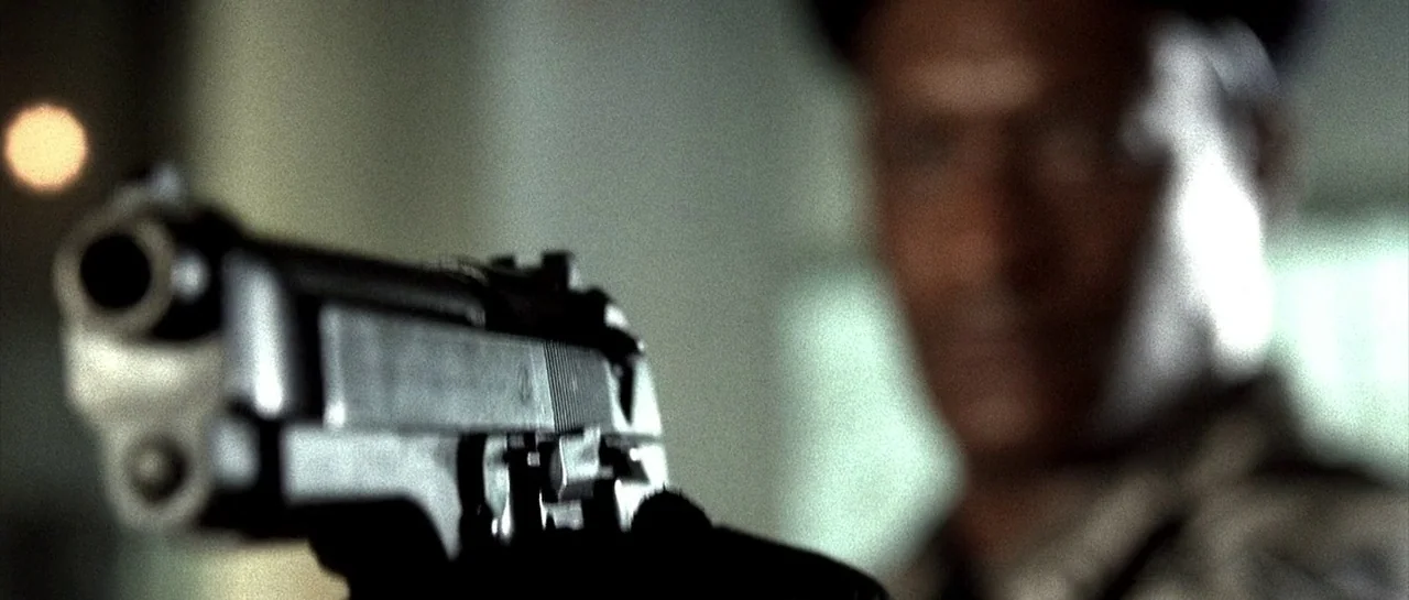 The Rock (1996) movie still: closeup — An interior space features a dark metal handgun held in the foreground. A person is visible in the blurred…