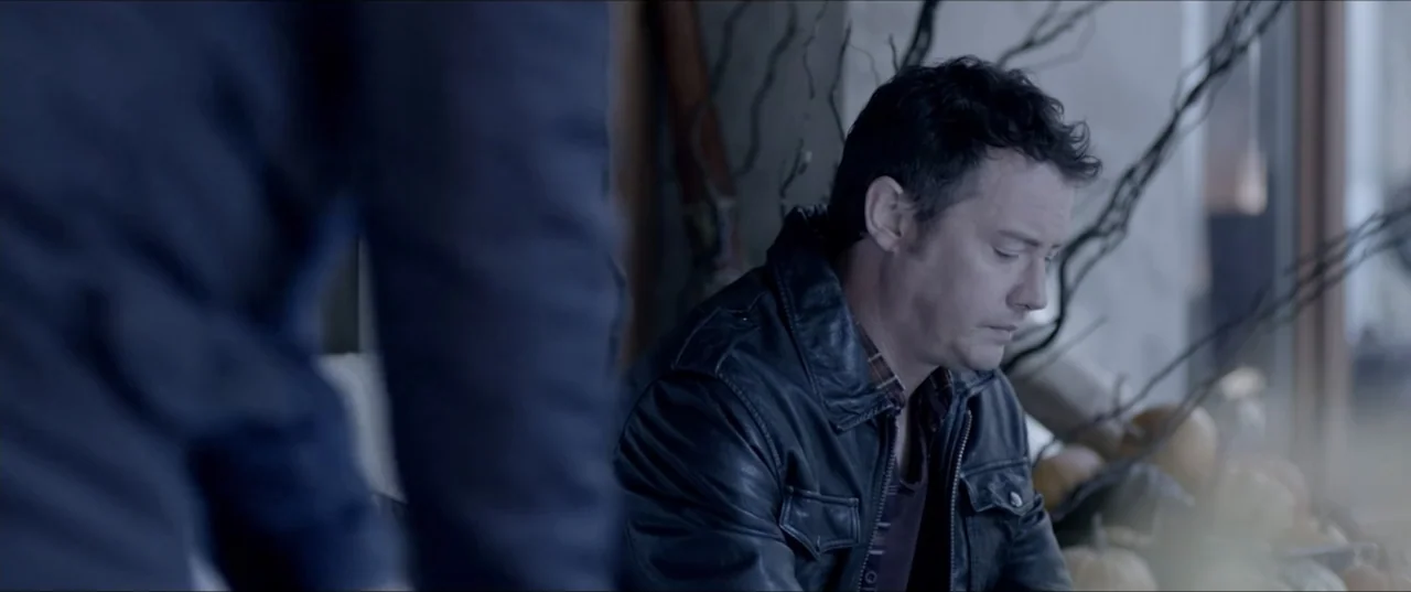Room 6 (2006) movie still: medium — An interior space features a man seated in a dark leather jacket near decorative branches. The man sits quietly…
