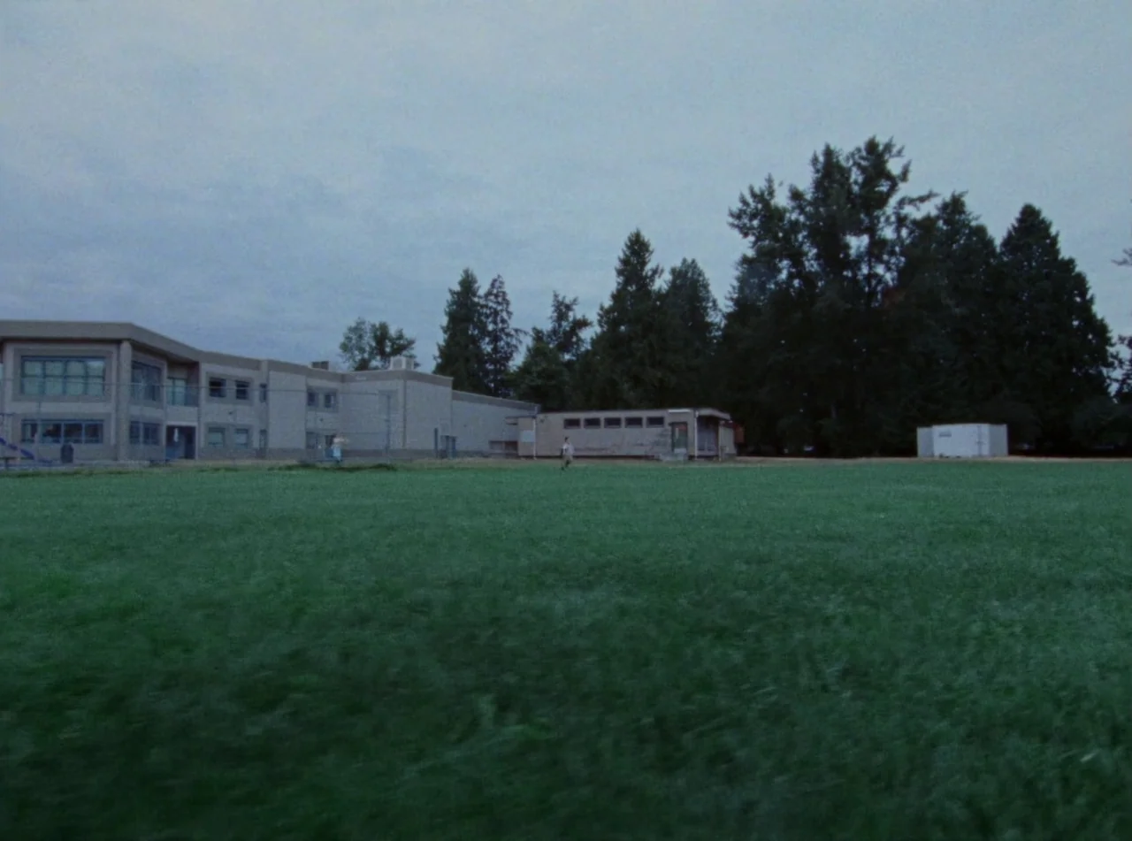 Riceboy Sleeps (2022) movie still: establishing — A wide, flat grass field stretches before a low, beige institutional school building. Tall…