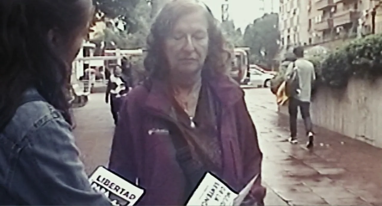 Belén (2025) movie still: medium — An urban sidewalk during the day features pedestrians in the background. A woman is being interviewed by an…