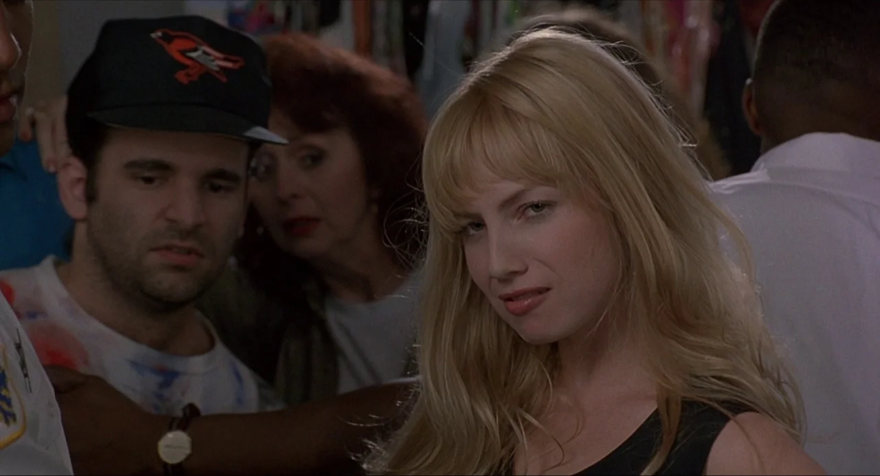 Serial Mom (1994) movie still: medium — A group of people stands in a retail space with clothing racks visible behind. A woman with blonde hair…