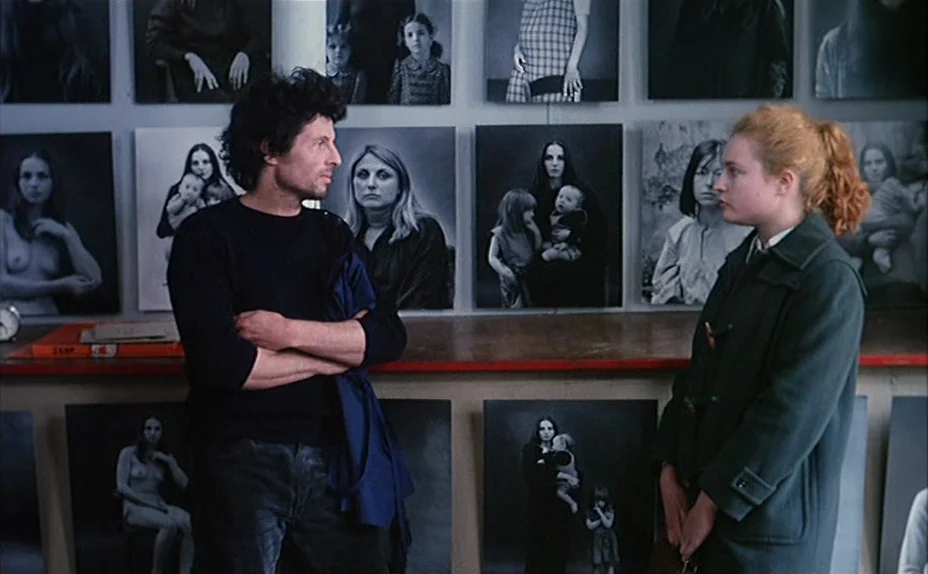 L’une Chante, l’Autre Pas (1977) movie still: medium — An interior space displays a wall covered in numerous framed black and white portraits. A man…