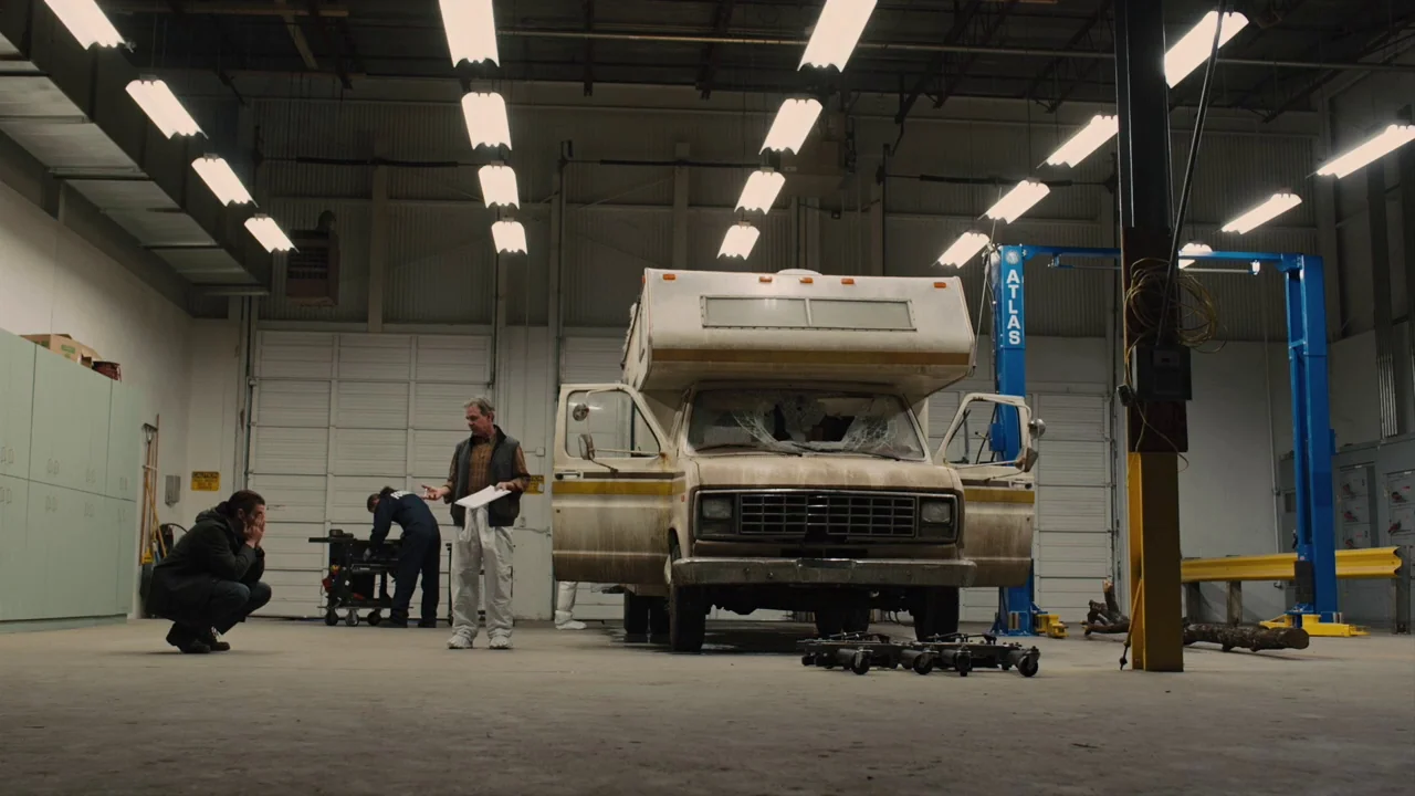 Prisoners (2013) movie still: wide — A spacious auto repair bay features concrete floors and bright fluorescent overhead lights. Several men stand…