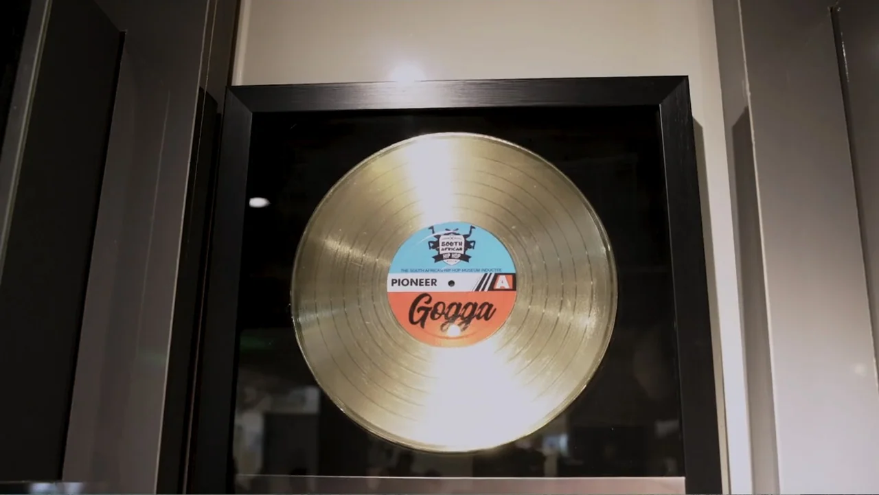African Throne World Tour (2024) movie still: closeup — A framed gold vinyl record is mounted on a plain interior wall. The record label features…