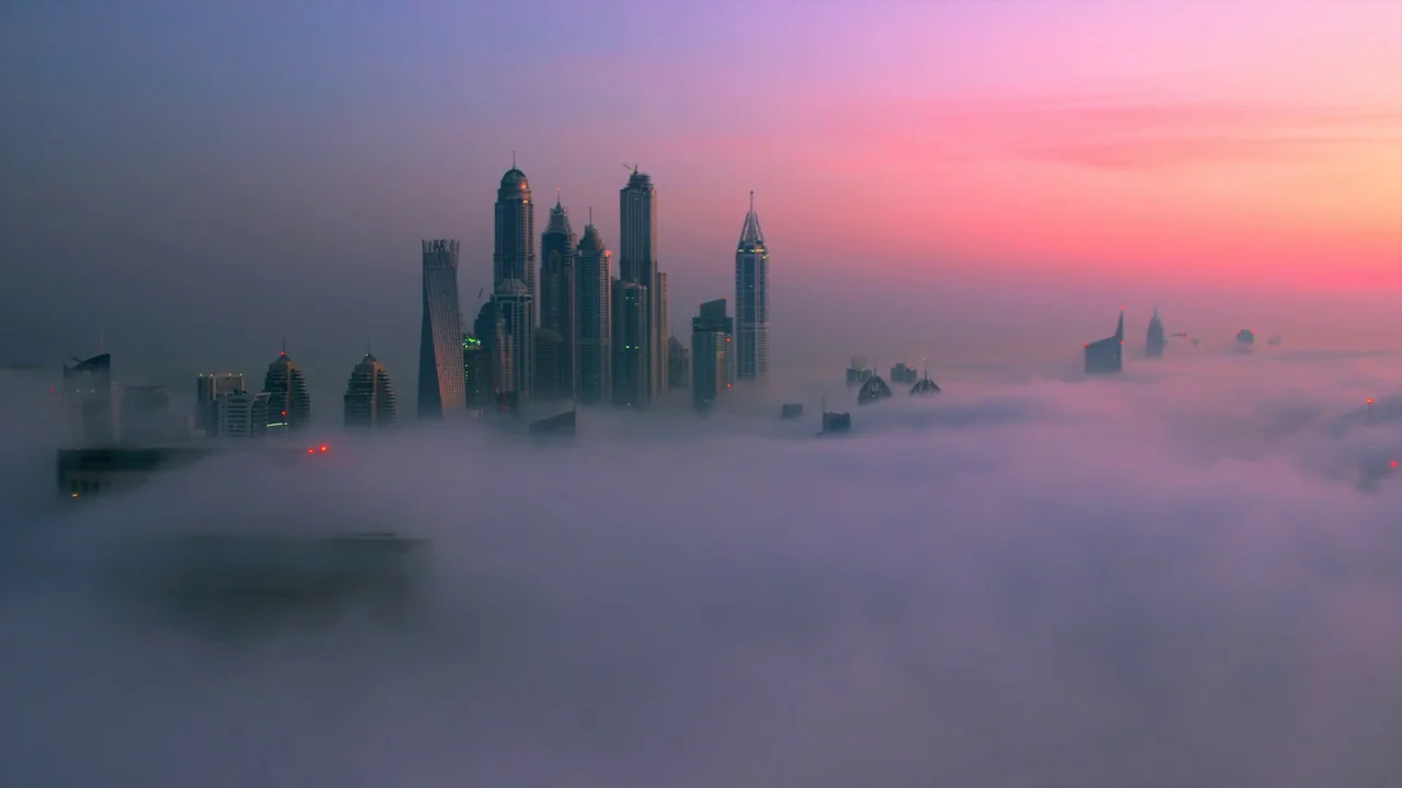 Awaken (2018) movie still: establishing — A dense blanket of morning fog rises around the tops of modern glass skyscrapers. The sun illuminates the…