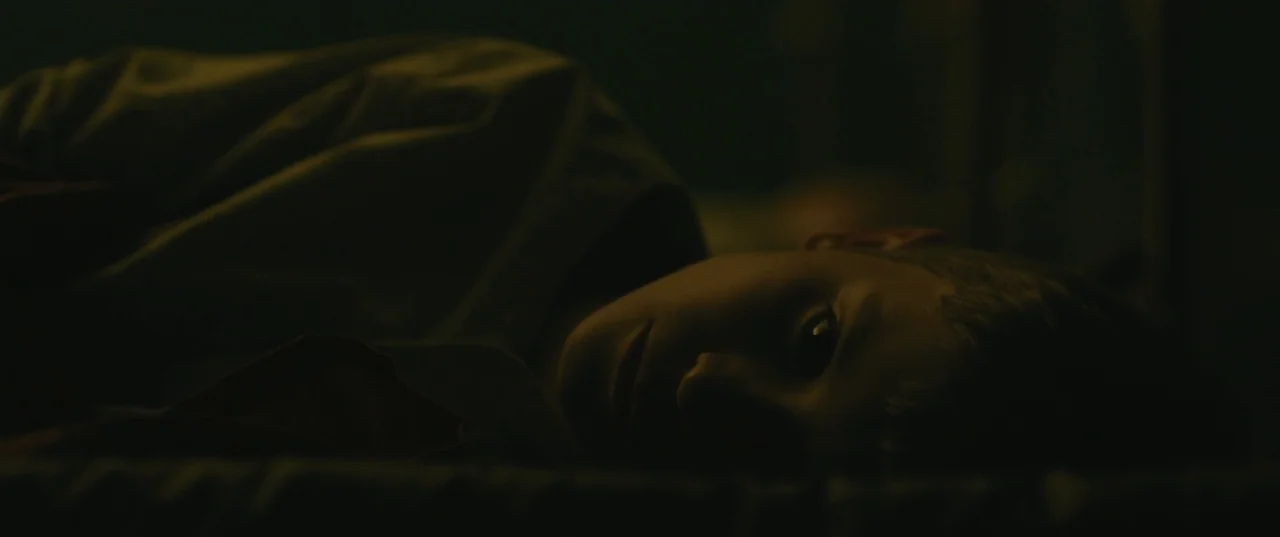 Evolution (2015) movie still: closeup — A dark bedroom environment with the child lying on a bed. The child gazes from the shadows toward the camera.