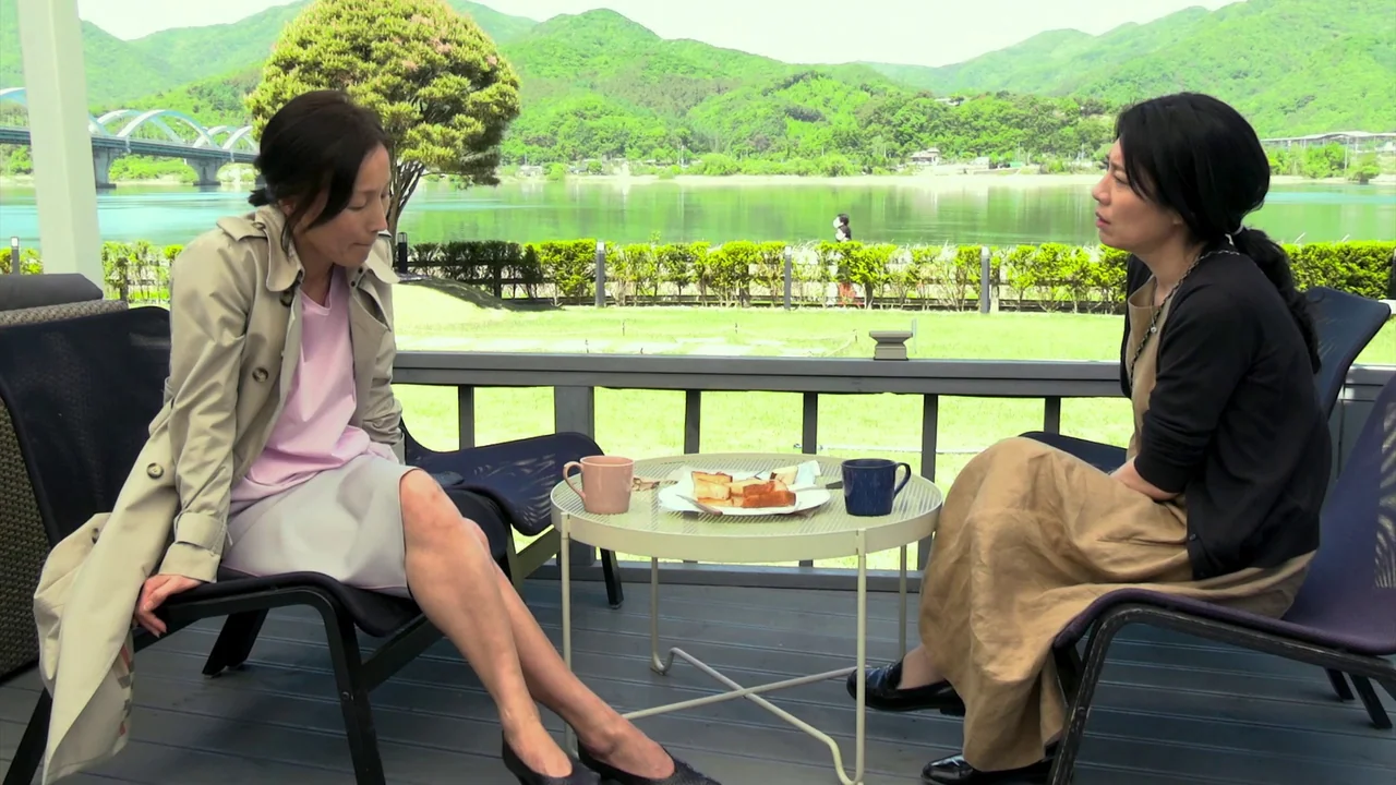 In Front Of Your Face (2021) movie still: fullbody — A wooden terrace overlooks a calm river with a bridge and green mountains beyond. Two women sit…