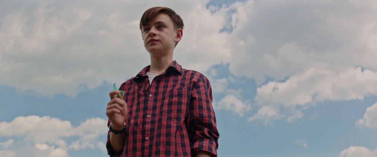It (2017) movie still: medium — A teenage boy stands in an open area under a bright, cloudy blue sky. He holds a small shard of green glass in his…