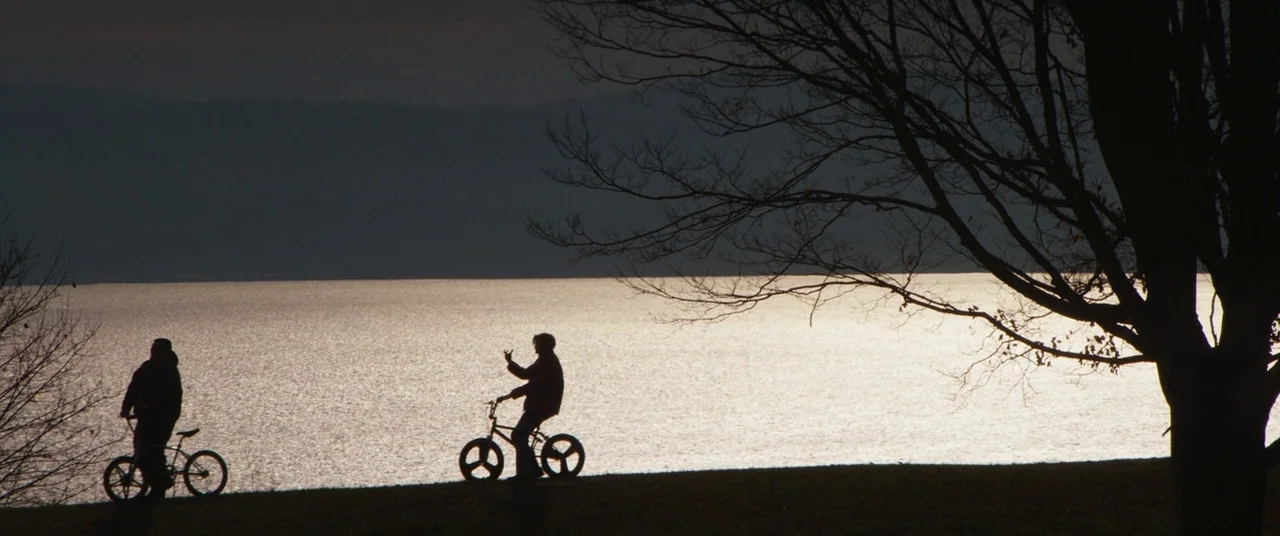 Super Dark Times (2017) movie still: wide — A grassy bank sits along a large body of water during the late afternoon. Two individuals ride bicycles…